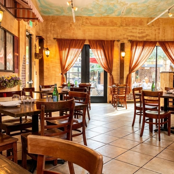 Cozy restaurant interior with wooden tables and chairs, warm lighting, and large windows with sheer curtains.