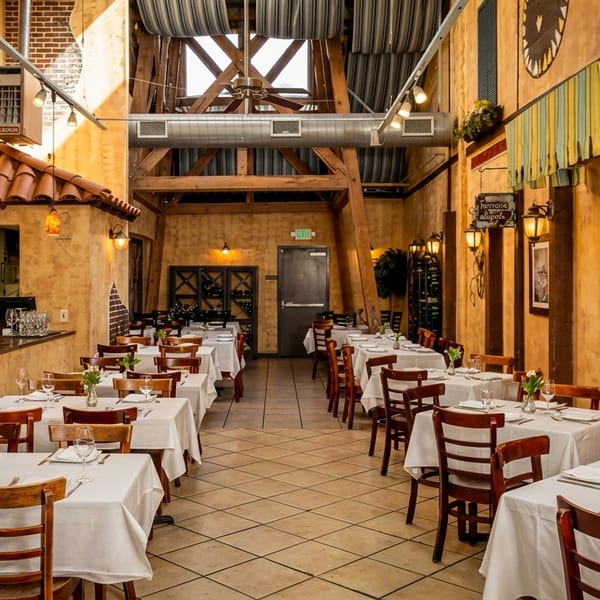 Warmly lit restaurant interior with white-clothed tables, wooden chairs, and a bar area under a high ceiling.