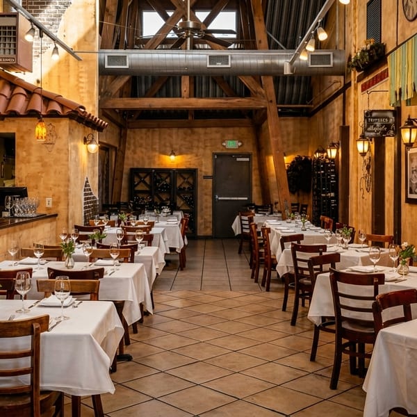 An empty Italian restaurant dining room with white-clothed tables, wooden chairs, and rustic decor.