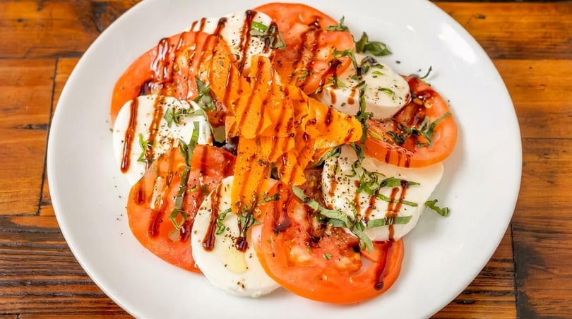 Caprese salad with sliced tomatoes, mozzarella, basil, balsamic glaze, and carrot ribbons on a white plate.
