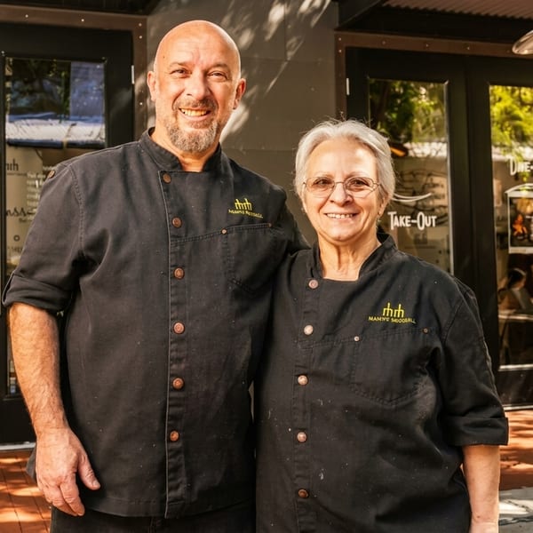 Two smiling chefs, a man and a woman, stand outside a restaurant entrance.