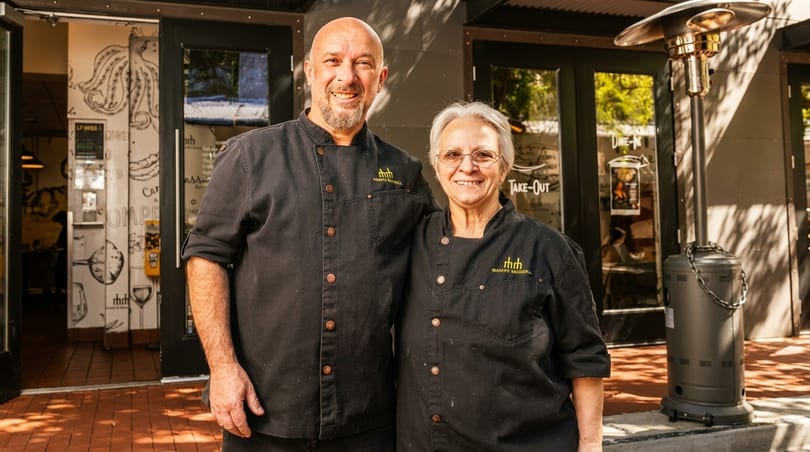 Two smiling chefs, a man and a woman, stand outside a restaurant entrance.
