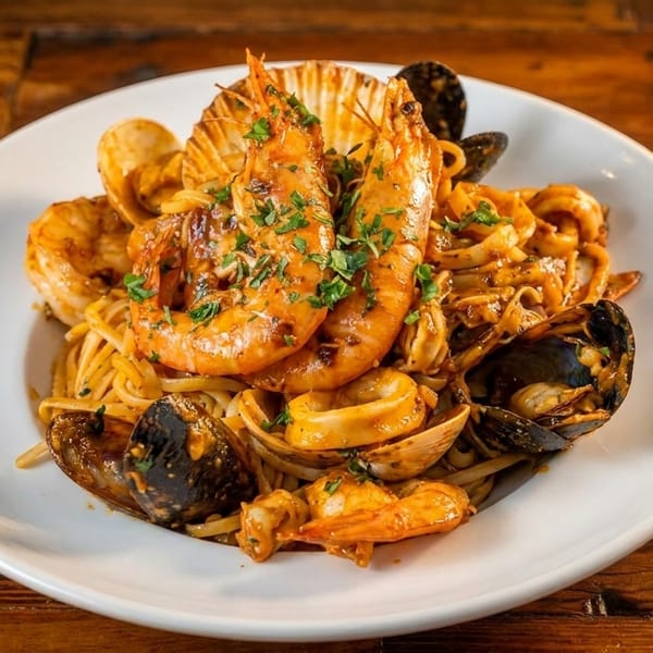 A white plate of seafood pasta with large shrimp, mussels, clams, and calamari on a wooden table.