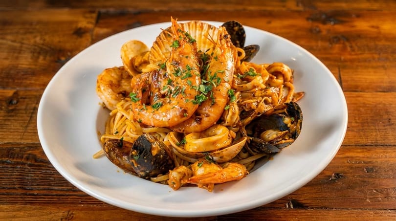 A white plate of seafood linguine with large shrimp, mussels, clams, and calamari on a wooden table.