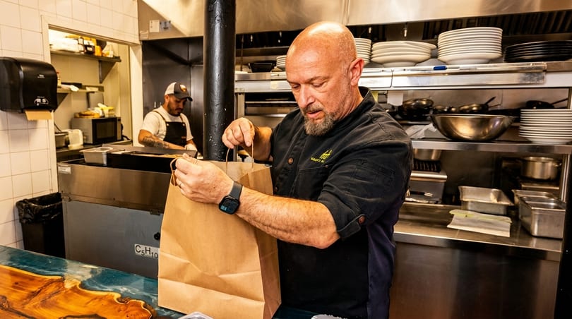 A chef in a black uniform carefully places items into a brown paper bag in a busy kitchen.
