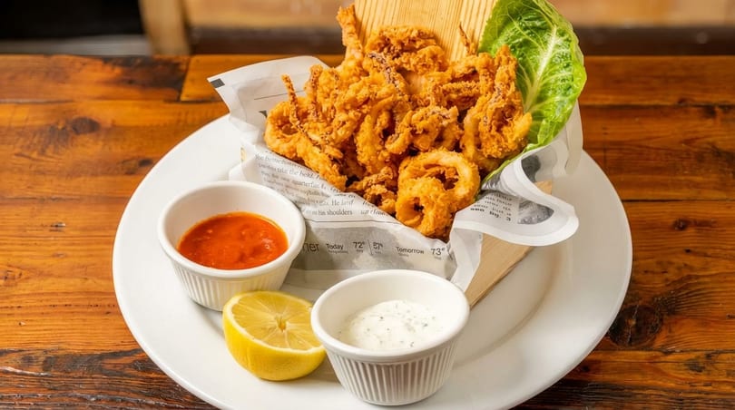 Crispy fried calamari served in a basket with lemon, marinara, and tartar sauces on a wooden table.