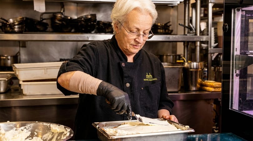 A chef in a black uniform with "Mamas Meatball" logo spreads white cream in a metal pan.